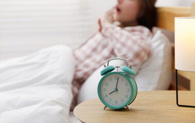 Young woman yawning on bed indoors, focus on alarm clock