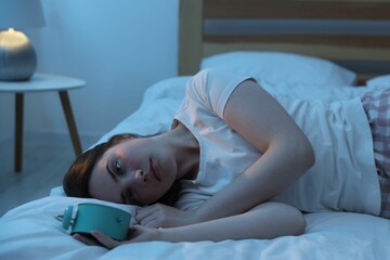 Frustrated young woman with alarm clock suffering from insomnia on bed indoors