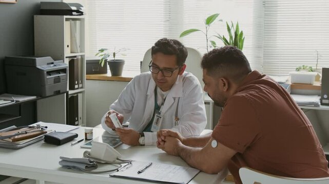 Male patient with glucose monitoring system patch on arm sitting in front of confident doctor presenting him new medication and listening to medical recommendations