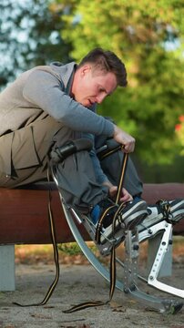Guy sitting on log in park, wearing gray long-sleeved shirt and cargo pants, focused on adjusting spring stilt straps attached to feet, surrounded by green trees, with low camera angle