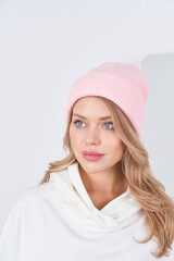Fototapeta premium Young woman with long hair wearing a pink knit beanie and a white cowl-neck top, looking to the side. Soft lighting enhances the serene and stylish appearance