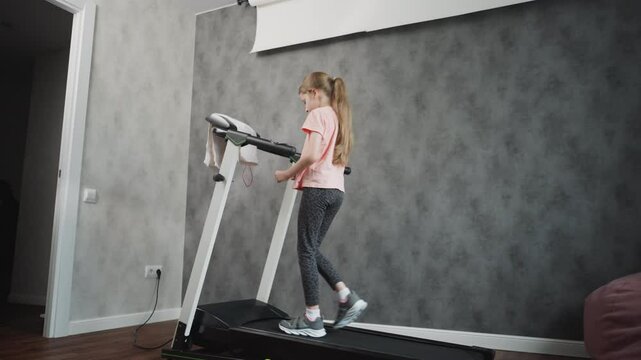 Rear view of young girl in patterned leggings walking on treadmill during home workout session long hair tied back hands on treadmill handles grey textured wall and wooden floor visible