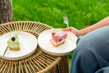 A woman is holding a plate of colorful layered cake while seated in a garden setting. The soft lighting and lush green background enhance the fresh and joyful mood of the scene.