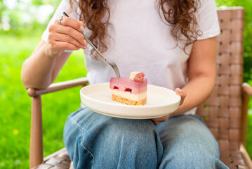 A woman is holding a plate of colorful layered cake while seated in a garden setting. The soft lighting and lush green background enhance the fresh and joyful mood of the scene.