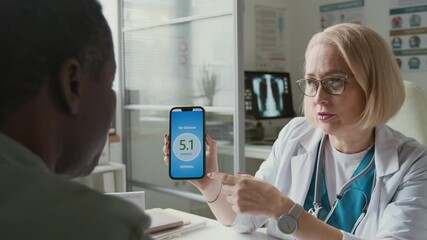 Confident mature female doctor sitting in front of male patient with diabetes and pointing at data on screen of smartphone in her hand during consultation in medical office - Powered by Adobe