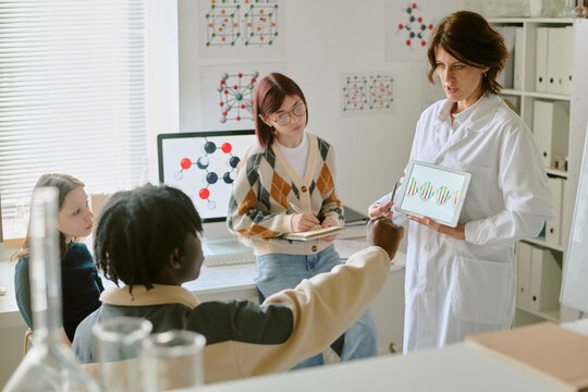 Woman in lab coat presenting data on tablet to diverse group of colleagues in office setting with charts and graphs on walls
