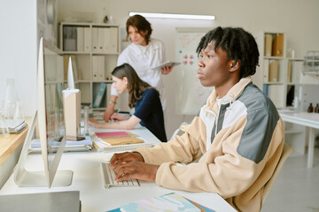 Student working on computer, focused and typing while two colleagues collaborate in background in...