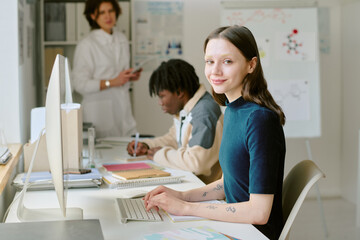 Obraz premium Portrait of student sitting at desk and smiling while classmates working in background in bright modern lab setting with computers and documents