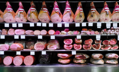 Shelves of a refrigerated display case stocked with various types of cured meats, including whole hams, sliced deli meats, sausages, salami, and mortadella.