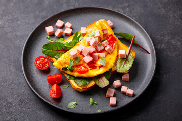 Toast with omelet, ham, cherry tomatoes and green salad on a black plate. Grey background. Close up.