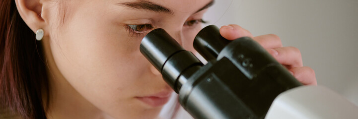 Young woman closely observing through microscope in lab setting. Capturing focused moment during scientific research, emphasizing concentration and attention to detail