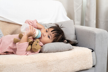 Asian toddler girl lying comfortably on beige sofa drinking milk from baby bottle while holding...