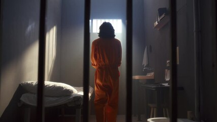 Young man in orange uniform walking around small sunlit prison cell, looking through barred window, then resting on bed - Powered by Adobe