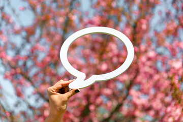 Hand holding a white speech bubble cutout with a backdrop of blooming pink cherry blossoms under a clear sky.