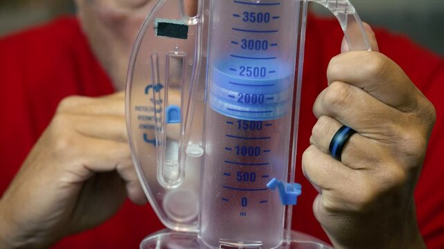 Close up view of a man using his incentive spirometer to measure lung capacity, demonstrating respiratory health recovery or breathing exercises in a home setting.  	