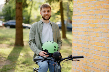 Young man smiles while holding a green helmet next to his bicycle in a sunny park during the...
