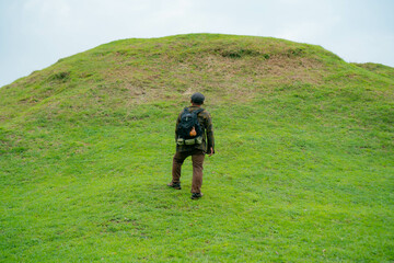 A male adventurer is enjoying the beauty of a small hill covered in green grass. The Asian male traveler is relaxing on the clean and beautiful green hills.
