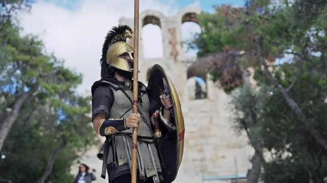 Confident hoplite spartan soldier wearing metal helmet and protecting with shield attacking with sharp spear with view of Acropolis Odeon of Herodes Atticus at background