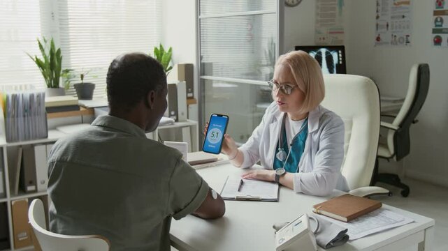 Doctor in lab coat sitting in front of male patient and scanning glucose monitor sensor on his arm with smartphone, then showing man data on screen and consulting him