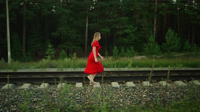 Side view of young lady in red dress running across railway sleeper while holding gown for balance, surrounded by gravel, grass, forest, and utility pole in background