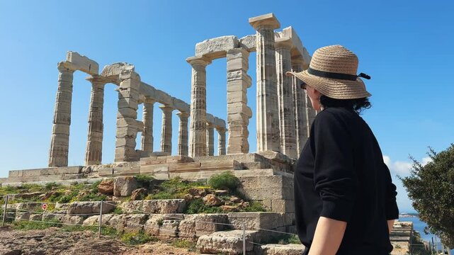 Woman tourist with hat and glasses sightseeing the Temple of Poseidon at Cape Sounion at the edge of Attica, Greece on summer sunny day