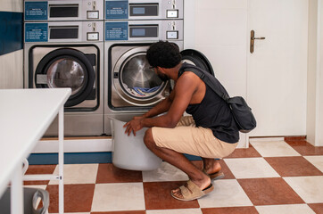 Young black man doing laundry in the laundromat, unloading the laundry from a plastic bin 