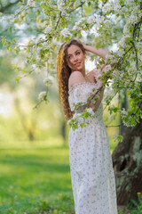 Beautiful Girl with long hair in a white dress poses near white blossoming apple trees