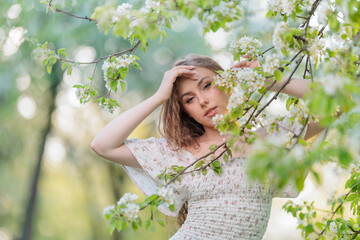 Beautiful Girl with long hair in a white dress poses near white blossoming apple trees