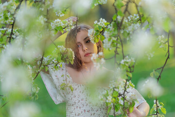 Beautiful Girl with long hair in a white dress poses near white blossoming apple trees