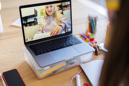 A student is video chatting with a classmate on her laptop. They are likely working on a school project together, as suggested by the notebook and art supplies nearby.