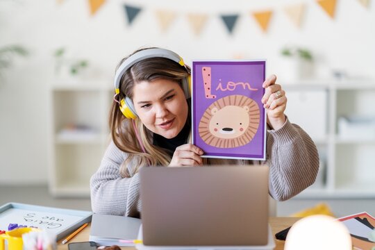 A teacher is leading a virtual lesson, showing a flashcard with a lion. She's teaching the alphabet to young children remotely.