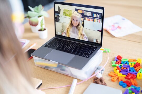 A student video calls her teacher for help with schoolwork. The laptop is on a box for better viewing. Colorful letters are on the desk, ready for learning.