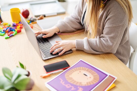 A woman is working on her laptop at a desk covered in children's learning materials. She's likely preparing lessons or activities for young kids.