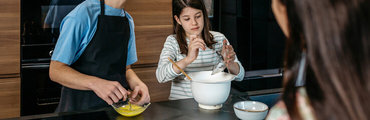 Banner of teenage boy and girl making a cake together in kitchen, whisking eggs and adding powdered sugar, while mother watching and following recipe on tablet. Family leisure time at home concept.