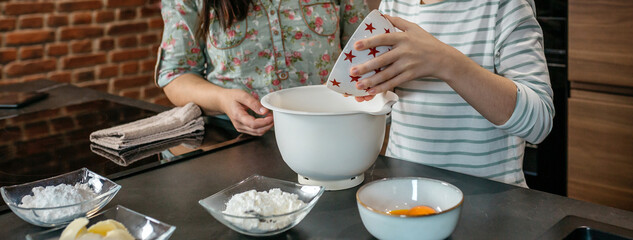 Unrecognizable young daughter pours flour into mixing bowl while baking with his mother in modern kitchen, surrounded by ingredients, sharing warm moment of culinary learning and family connection.