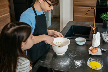 Boy and young girl preparing together homemade cake, mixing ingredients in bowl and sharing culinary learning experience with joy and teamwork. Brother and sister baking cake in modern kitchen at home