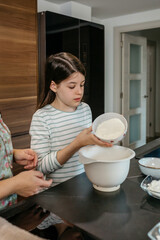Concentrated teenage girl pours flour into a mixing bowl while baking in a modern kitchen surrounded by various ingredients. Young female child learning cooking skills at home concept.