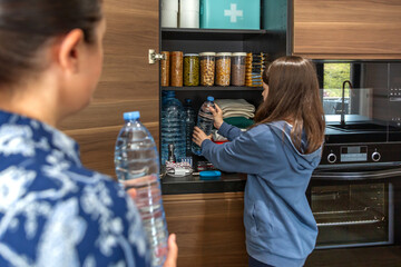 Girl storing water bottles in kitchen pantry with food and first aid kit ready for war or natural disasters. Family preparing 72 hour survival kit as part of foresight and self sufficiency strategy.