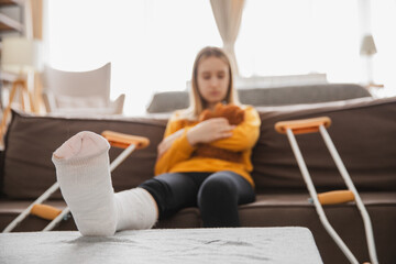 Broken leg. Young girl with broken leg cast sitting on sofa. Sad little kid recovering from broken...