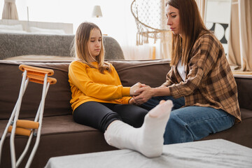 Girl with broken leg in cast sitting with mother on couch at home. Sad teenage kid recovering from broken leg. Girl using cast crutches for quick rehabilitation. Mom supporting daughter
