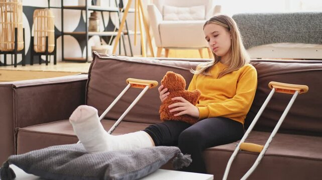 Young girl with broken leg cast sitting on sofa. Sad little kid hugging her teddy bear recovering from broken leg at home. Girl using cast and crutches. Accident injury fracture treatment foot bandage