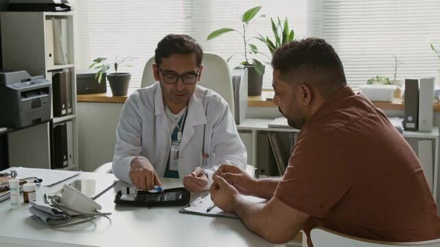 Young confident male clinician sitting by workplace in front of Hispanic man, looking at him and giving instructions about usage of glucose level monitoring set