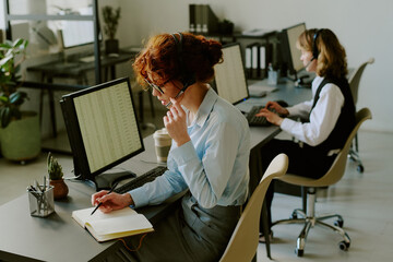 Obraz premium Two office workers focused on tasks at their desks, with one taking notes and wearing headset. Modern workspace with computers and plants creating productive environment