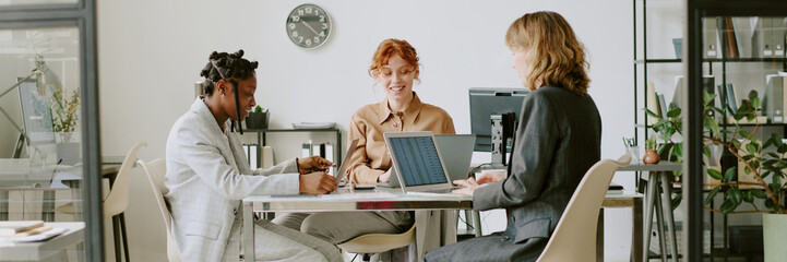 Three professionals engaged in collaborative meeting with laptops and documents on table. Diverse group working on project in spacious, well-lit office