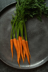Fresh raw and peeled carrots, with the green still attached, lie in a large metal bowl. The bowl is gray. The carrots glow orange.