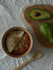 White linen tablecloth with bowl of oatmeal and tomato paste, topped with fried egg. Wooden spoon, cutting board with avocado half and slices. Natural light, cozy atmosphere.