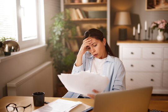 A woman with a concerned expression examines multiple papers related to insurance problems in her well-lit home office. She sits at a wooden desk cluttered with documents and a laptop.