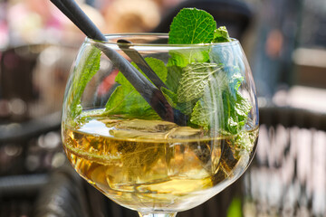 Cold summer drink or mocktail in a glass with ice cubes, fresh green mint leaves and a straw on a terrace in summer.