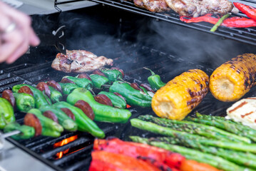 Grilling chicken, corn cobs, paprika, padron, asparagus with temperature control. Meat tongs in human hands.