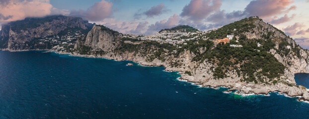 Capri Island's rugged cliffs meet the Tyrrhenian Sea, with whitewashed villas on green hillsides under a vibrant sky with soft clouds. © Aerial Film Studio